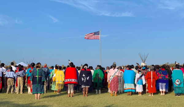 Members of the Kiowa Gourd Clan Ceremony stand as the flag of Spencer “Corky” Sahmaunt is raised. Carnegie, Oklahoma; July 4, 2019. Mr. Sahmaunt served in the U.S. Army during the Korean War and was a member of the Kiowa Black Leggings Warrior Society, as well as the Kiowa Gourd Clan.The Kiowa Flag Song, analogous to the Star Spangled Banner, accompanied the flag-raising. (Photo courtesy of Mari Frances Sahmaunt, used with permission)